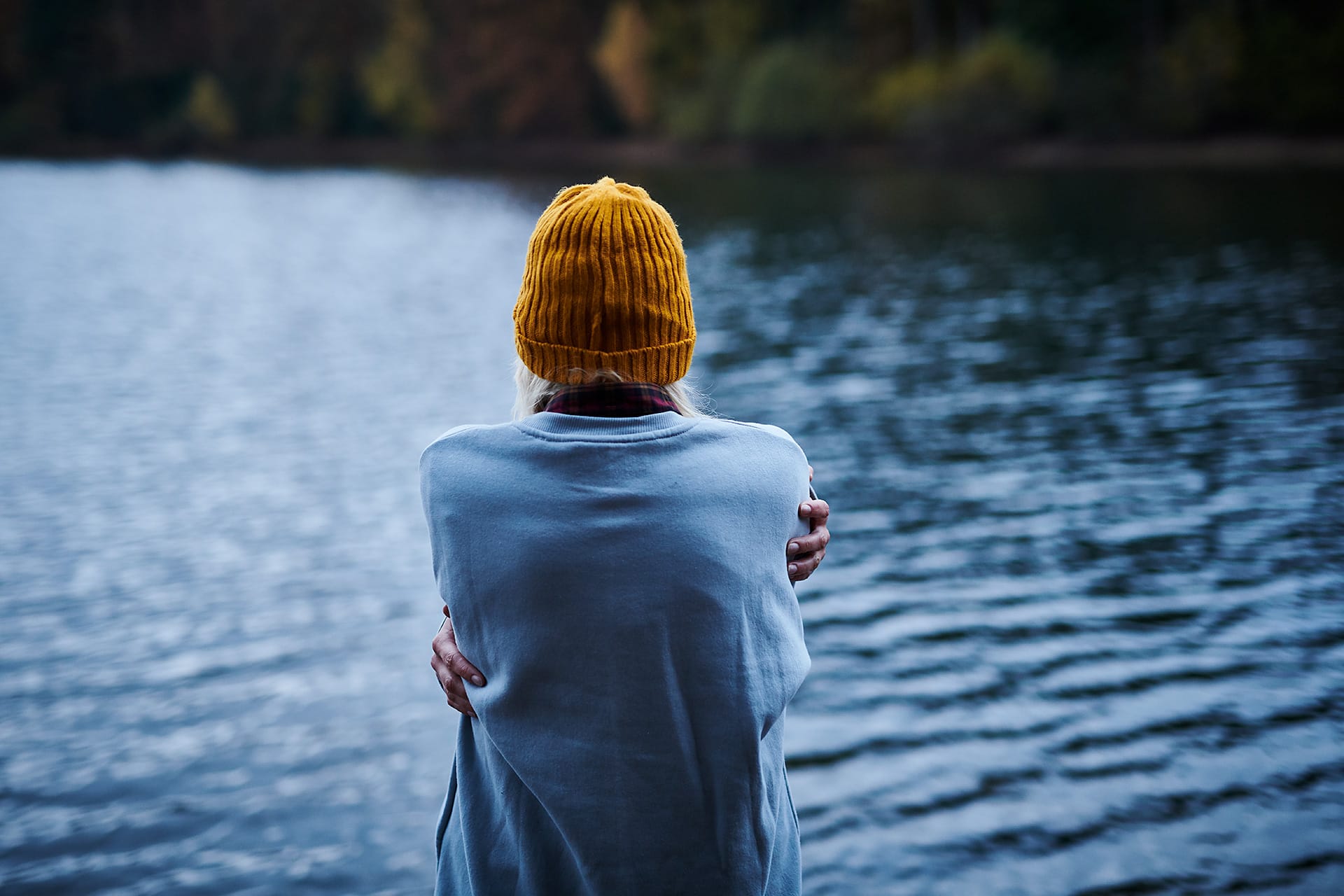 Junge Frau umarmt sich selbst vor einem See. Rückansicht