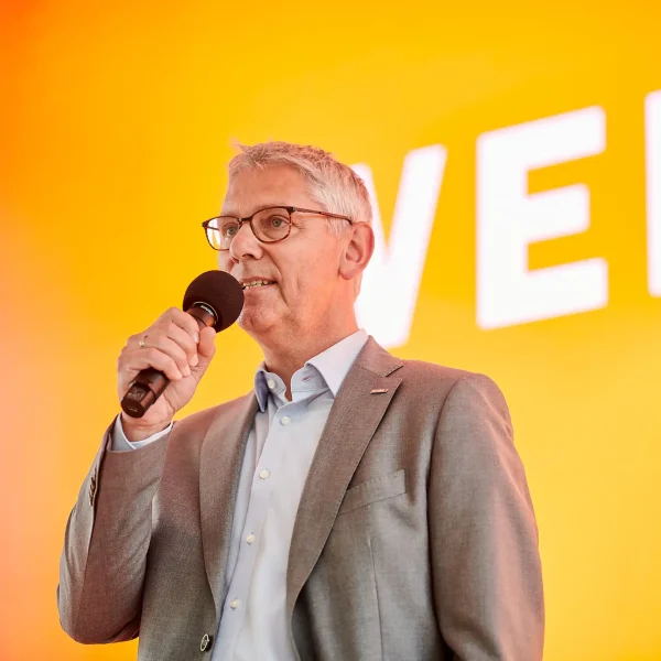 Redner mit Mikrofon und Brille spricht vor orangefarbener Bühnenprojektion bei Eventfotografie in Dortmund.